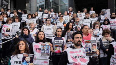 Viele Menschen stehen auf einer Treppe und tragen Warnwesten mit dem Aufdruck "Press". Einige Warnwesten haben rote Flecken. Die Menschen halten Plakate mit Bildern von verschiedenen Journalisten hoch.